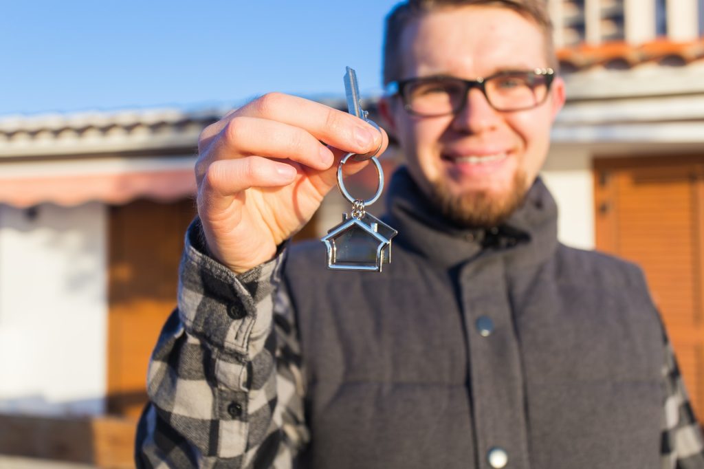 Dwelling, buying home, real estate and ownership concept - handsome man showing his key to new home